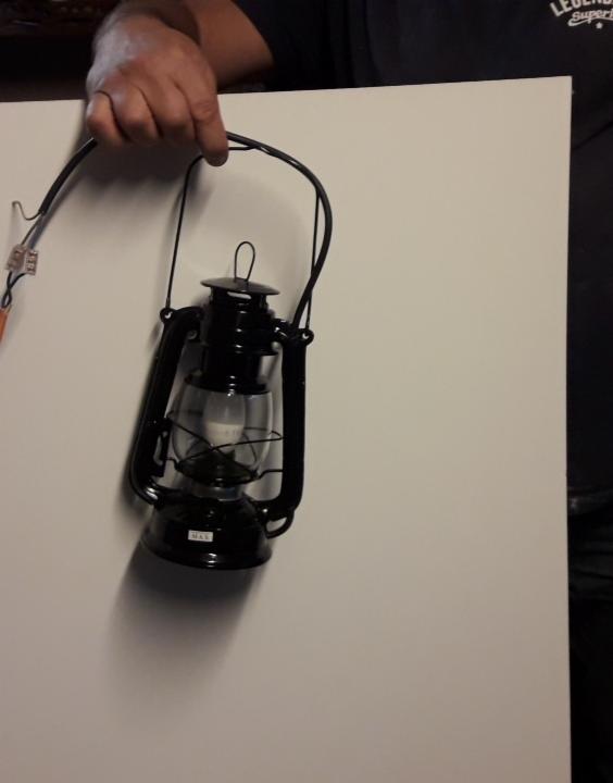 Czarna lampa naftowa z kablem elektrycznym, trzymana przed białą ścianą. Widoczne złącza kablowe.