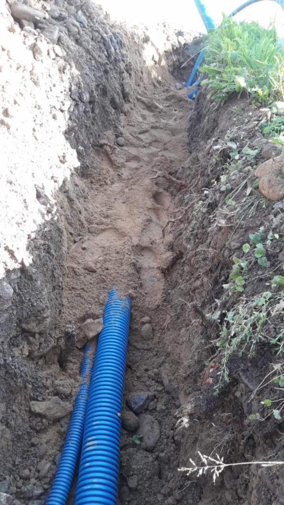 Wykop z ułożonymi niebieskimi rurami karbowanymi, widoczne ślady stóp na dnie wykopu i roślinność na krawędzi.