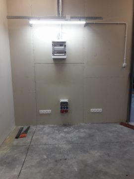 Szara ściana z zamontowaną skrzynką rozdzielczą, gniazdami elektrycznymi i listwą oświetleniową LED. Widoczne fragmenty narzędzi na podłodze.