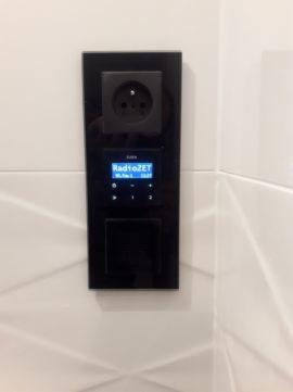 Czarny panel sterowania z gniazdkiem elektrycznym i wyświetlaczem radia wbudowany w białą, wzorzystą ścianę z płytek.