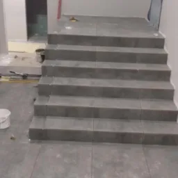 Wyłożone szarymi płytkami schody w trakcie prac wykończeniowych; widoczna także podłoga z płytek, narzędzia glazurnicze oraz wiadro z klejem.