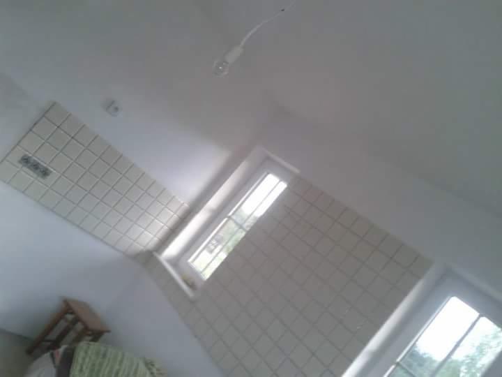 Niedokończona kuchnia z białymi płytkami na ścianie, widoczne okno i wisząca żarówka, ujęcie pod kątem.