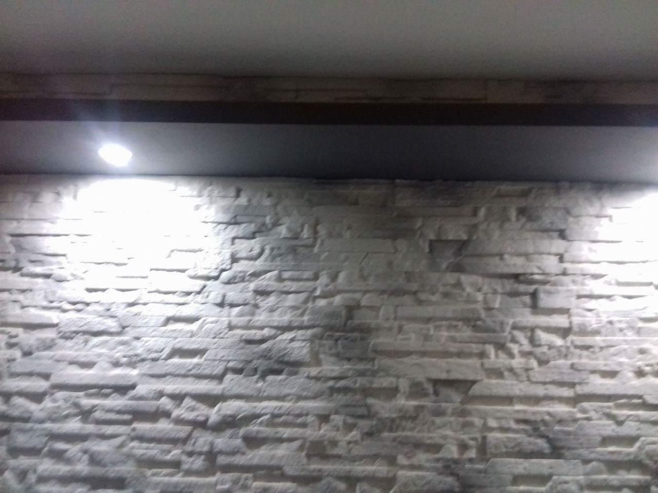 Ściana wykończona dekoracyjnym kamieniem w odcieniach bieli i szarości, oświetlona punktowo od góry, z widoczną ciemną półką lub listwą pod sufitem.