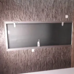 Szara ściana z dekoracyjną okładziną imitującą kamień, z zamontowanym lustrem w srebrnej ramie i widocznymi elementami instalacji elektrycznej.