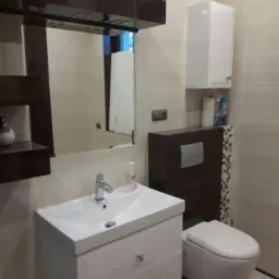 Nowoczesna łazienka z białą umywalką z szafką, lustrem z ciemnobrązową półką, wiszącą szafką i toaletą w zabudowie.