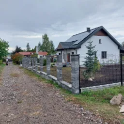 Nowoczesny dom jednorodzinny z panelami słonecznymi i częściowo ukończonym ogrodzeniem z betonowych słupków i metalowej siatki, widok z szutrowej drogi.