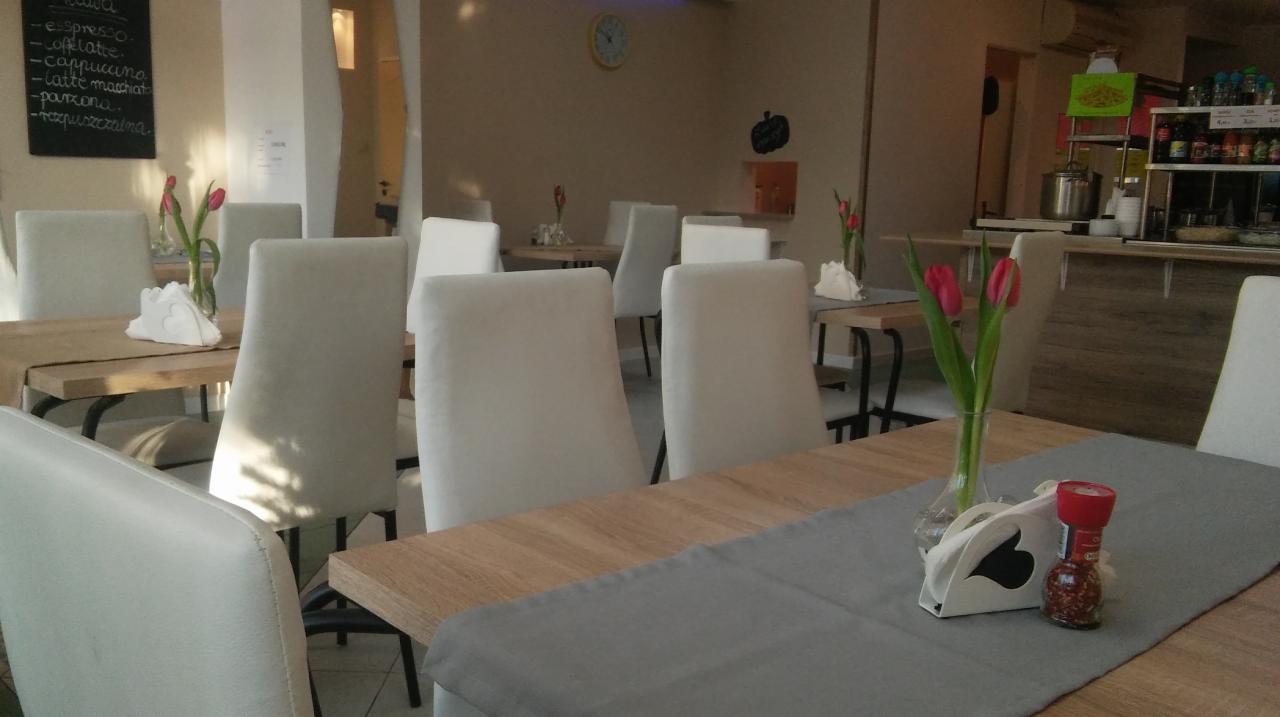 Jasne wnętrze restauracji z drewnianymi stolami nakrytymi szarymi bieżnikami, białymi krzesłami, tulipanami w wazonach i menu wypisanym kredą na tablicy.
