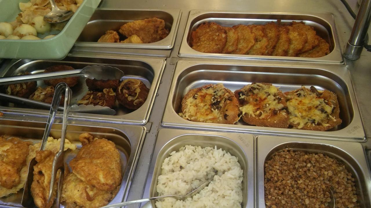 Bufet z różnorodnymi daniami: kotlety z cebulą, placki ziemniaczane, zapiekanki z serem, ryż biały i kasza gryczana, prezentowane w metalowych pojemnikach.