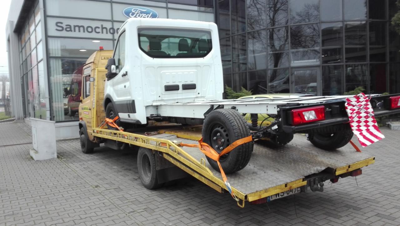 Żółta laweta z białym podwoziem samochodu na platformie, zabezpieczonym pomarańczowymi pasami transportowymi, stoi na tle salonu samochodowego Ford w Łodzi.