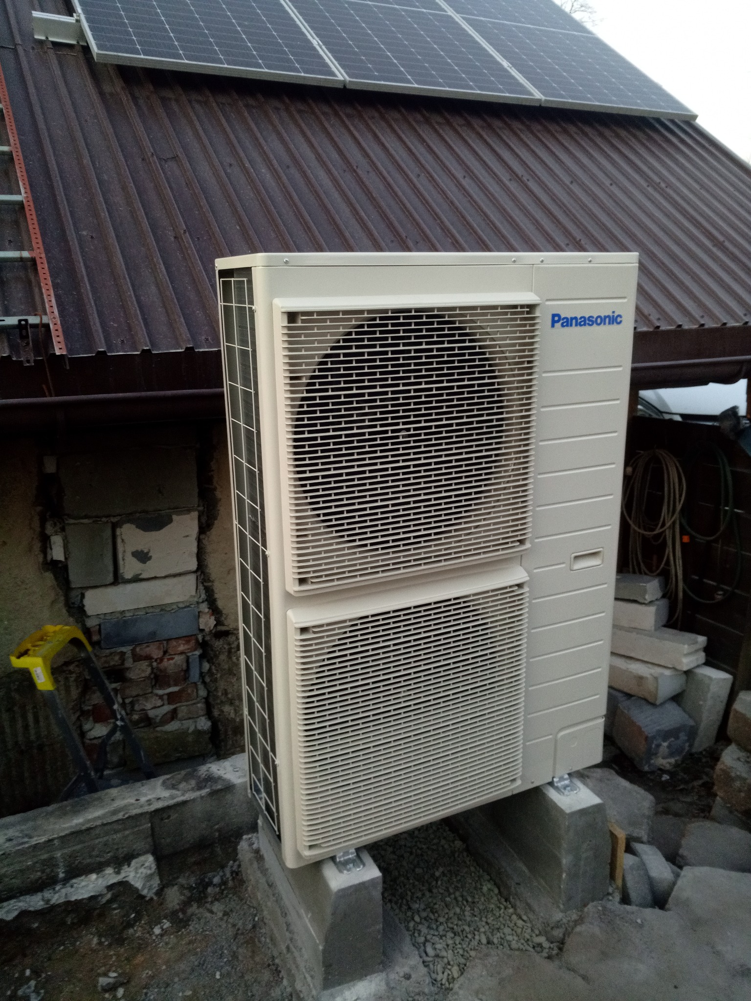 Zewnętrzna jednostka pompy ciepła Panasonic T-CAP 9 KW, zamontowana na betonowych podstawach przy budynku z panelami słonecznymi na dachu.