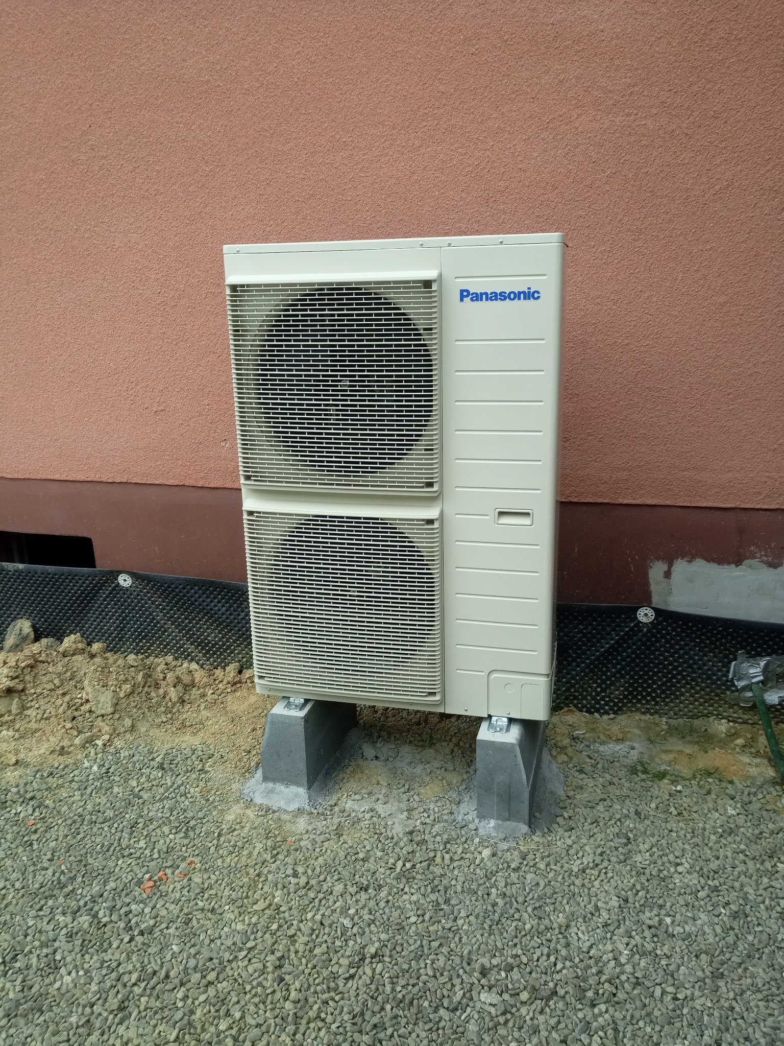 Zewnętrzna jednostka pompy ciepła Panasonic T-CAP 9 KW zamontowana na betonowych podstawach przy ścianie budynku z widoczną izolacją fundamentów i żwirowym podłożem.