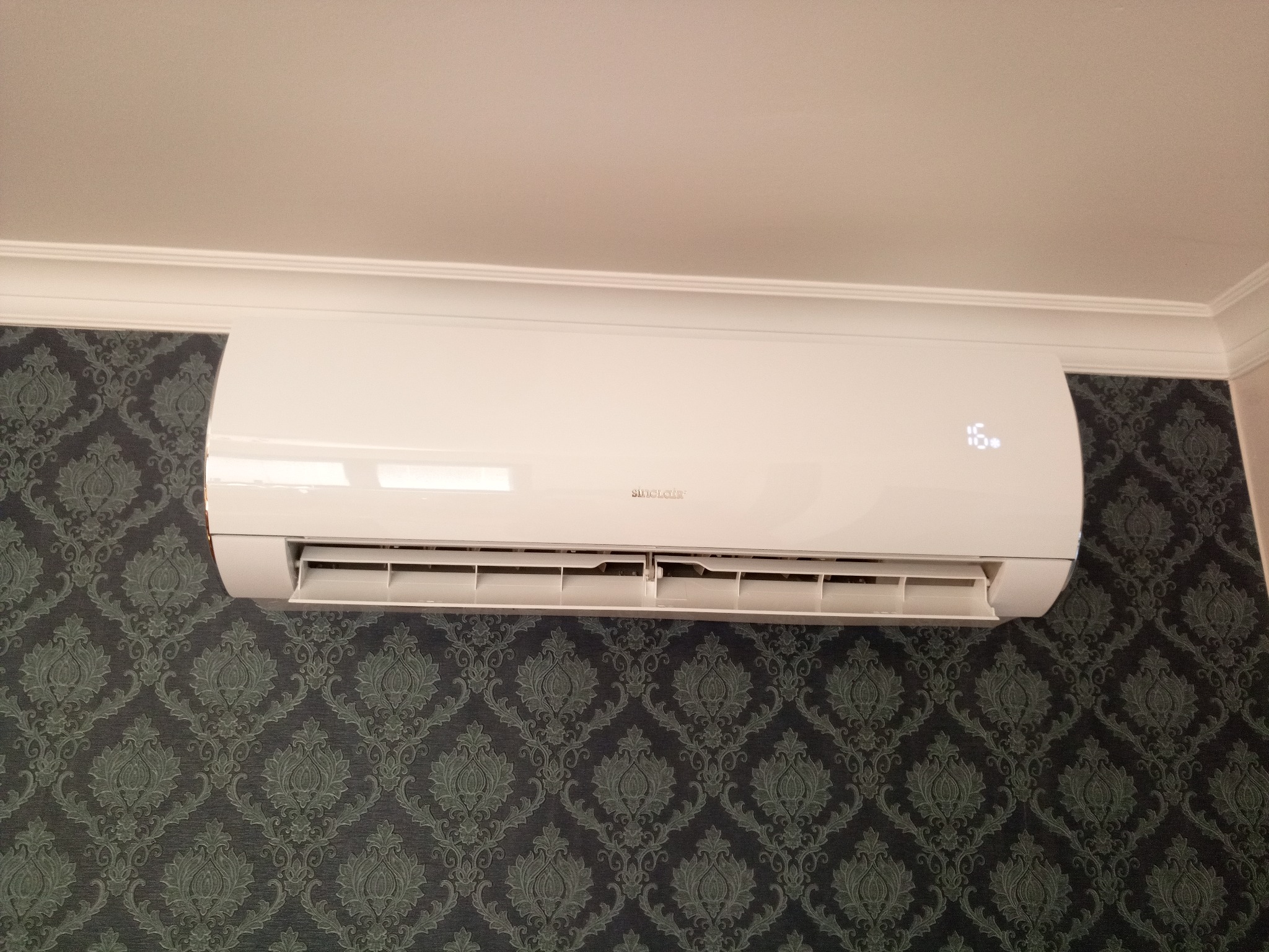 Biała jednostka wewnętrzna klimatyzacji marki Sinclair zamontowana na ścianie z ciemnozieloną tapetą w ornamenty, widoczny wyświetlacz temperatury 16 stopni.