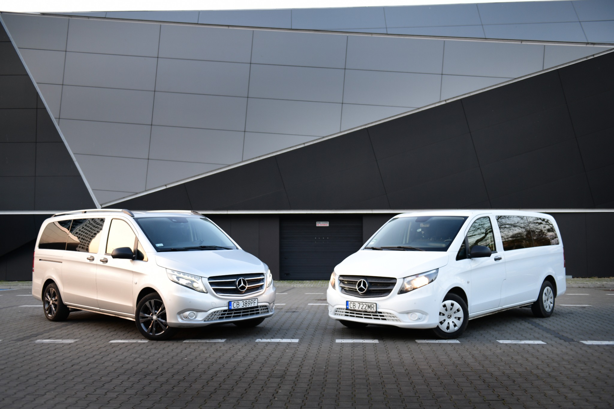 Dwa busy Mercedes Vito, srebrny i biały, zaparkowane naprzeciwko nowoczesnego budynku z geometryczną fasadą.