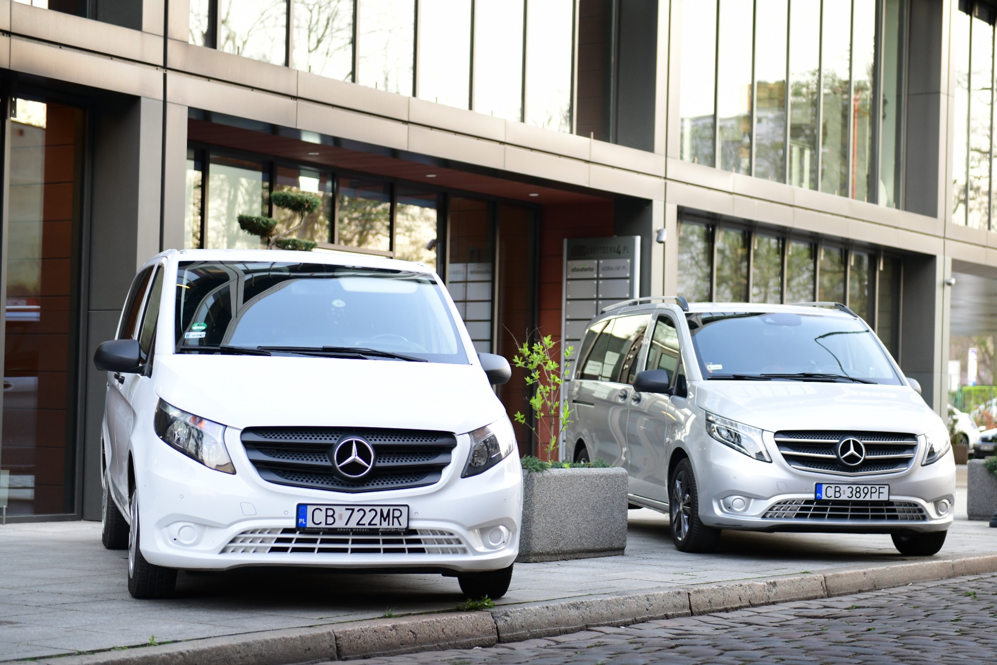 Dwa białe i srebrne busy Mercedes Vito zaparkowane przed nowoczesnym budynkiem biurowym z dużymi oknami, widoczne tablice rejestracyjne CB 722MR i CB 389PF.