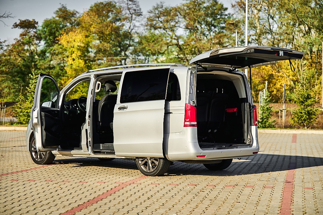 Srebrny van z otwartymi drzwiami przesuwnymi i klapą bagażnika na parkingu z kostki brukowej, jesienne drzewa w tle.
