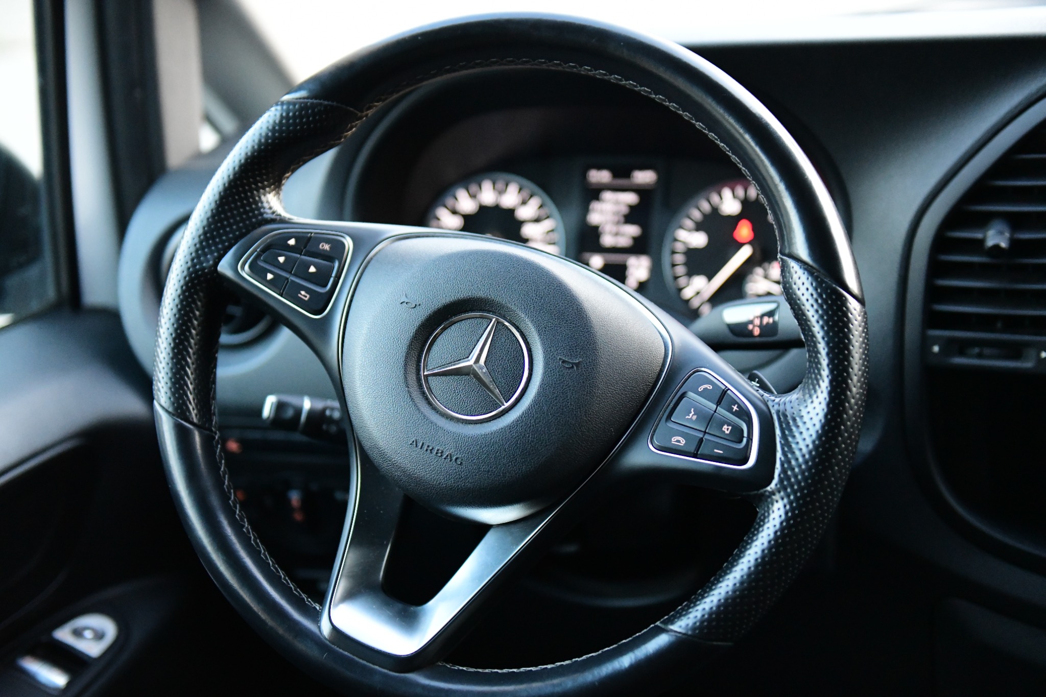 Czarna, skórzana kierownica z logo Mercedesa, widoczne przyciski sterowania, zegary i kratka wentylacyjna w tle.