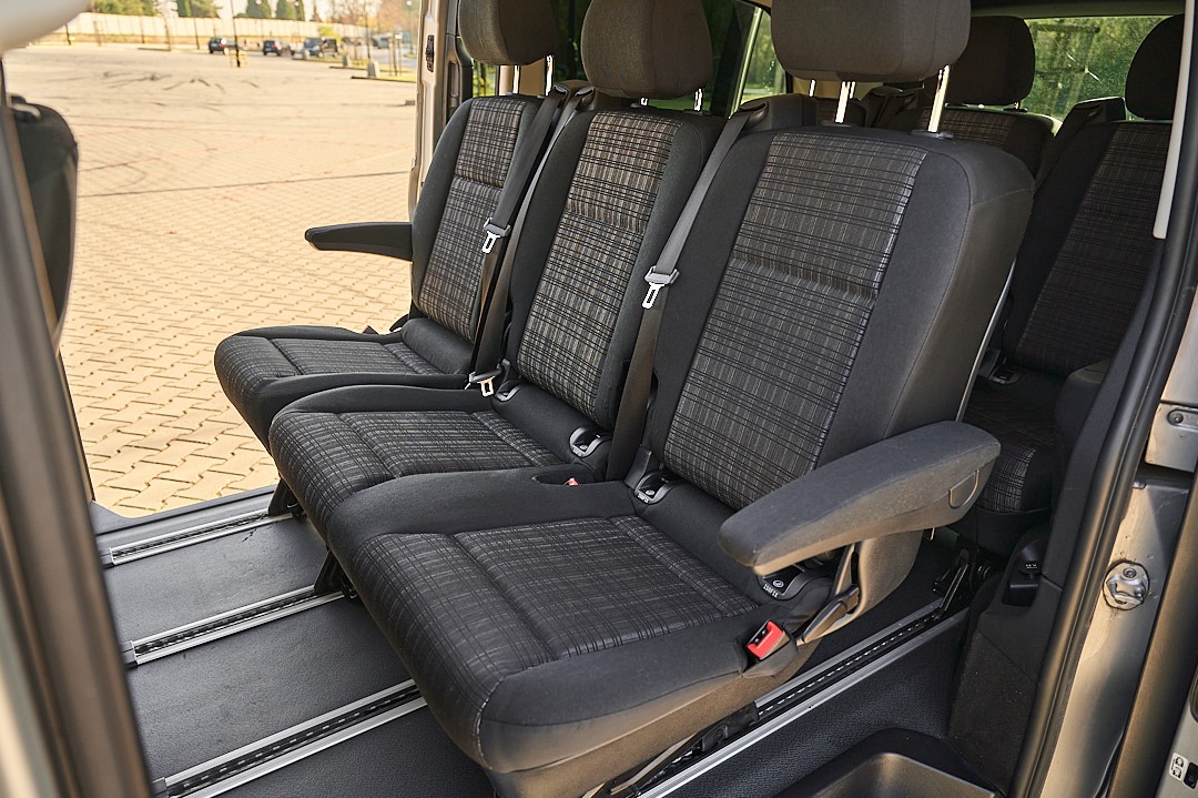 Wnętrze busa z trzema rzędami siedzeń z czarno-szarą tapicerką w kratę, widoczne szyny mocujące fotele.