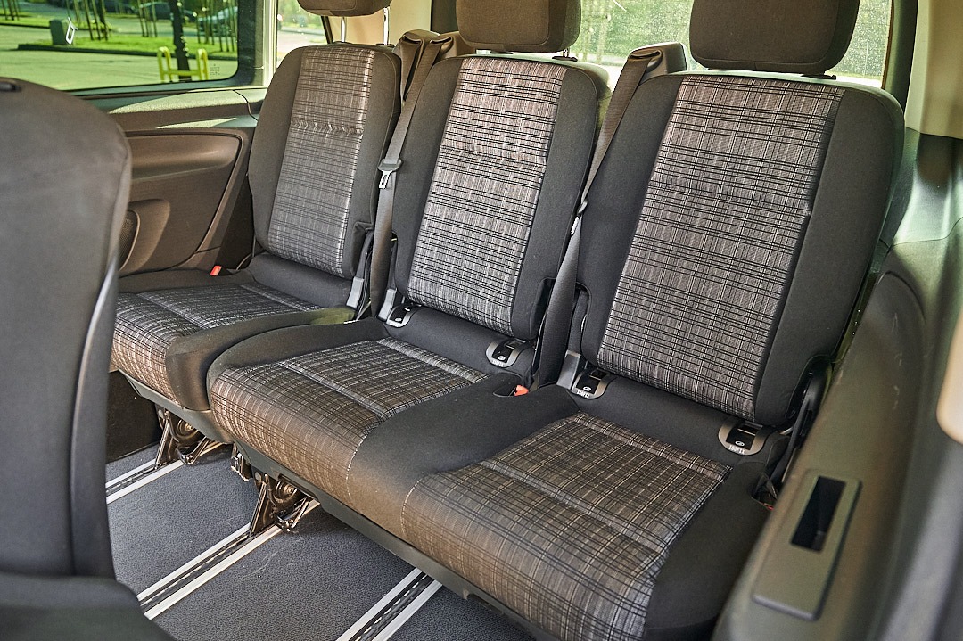 Wnętrze busa z trzema siedzeniami z szaro-czarną tapicerką w kratę, widoczne szyny podłogowe.