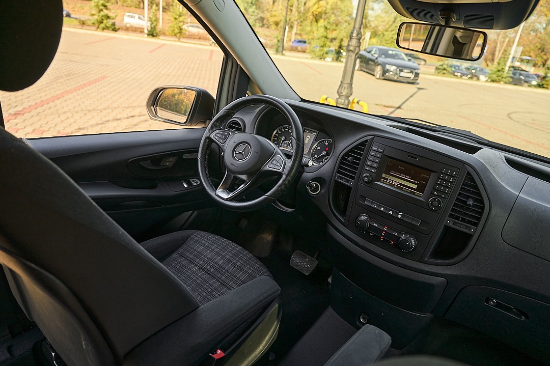 Wnętrze busa marki Mercedes-Benz Vito z widoczną deską rozdzielczą, kierownicą z logo Mercedes oraz fotelami z tapicerką w kratę.