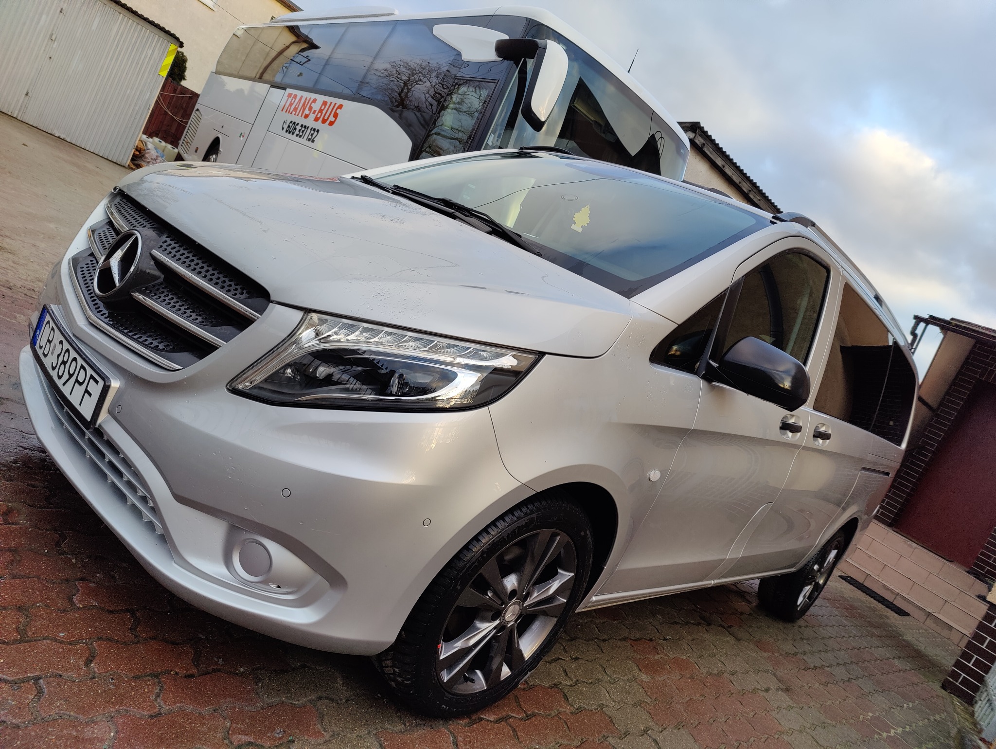 Srebrny Mercedes Vito z czarnymi felgami na tle białego autokaru TRANS-BUS, zaparkowany na czerwonej kostce brukowej.