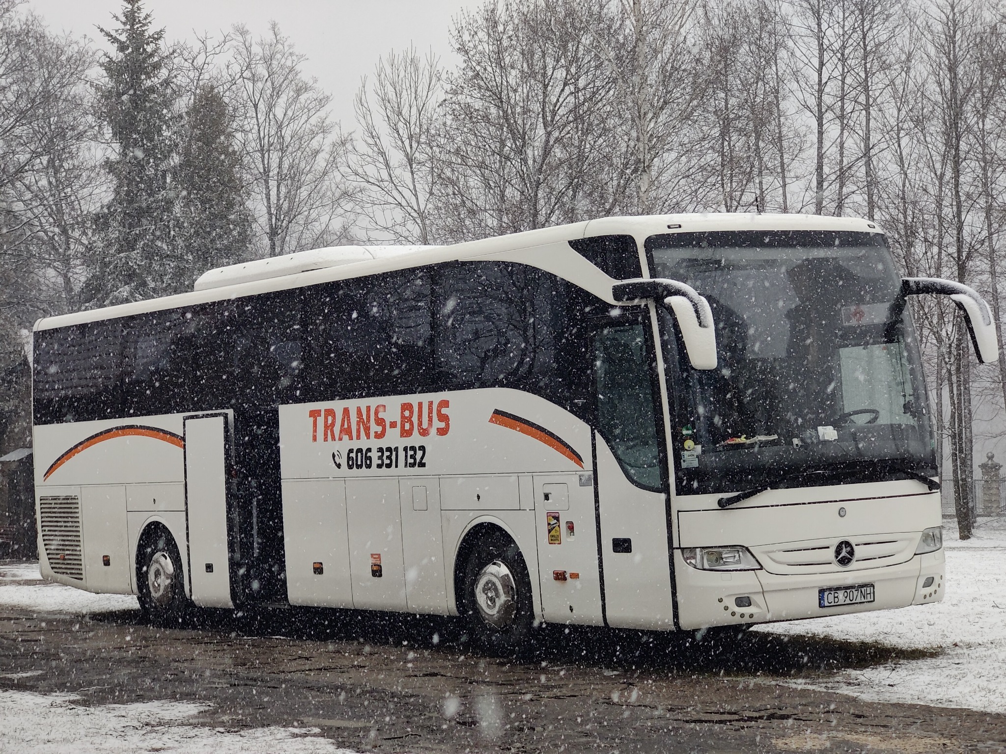 Biały autokar marki Mercedes z otwartymi drzwiami, oznaczony logo 'TRANS-BUS' i numerem telefonu, stoi na śniegu w zimowy dzień.