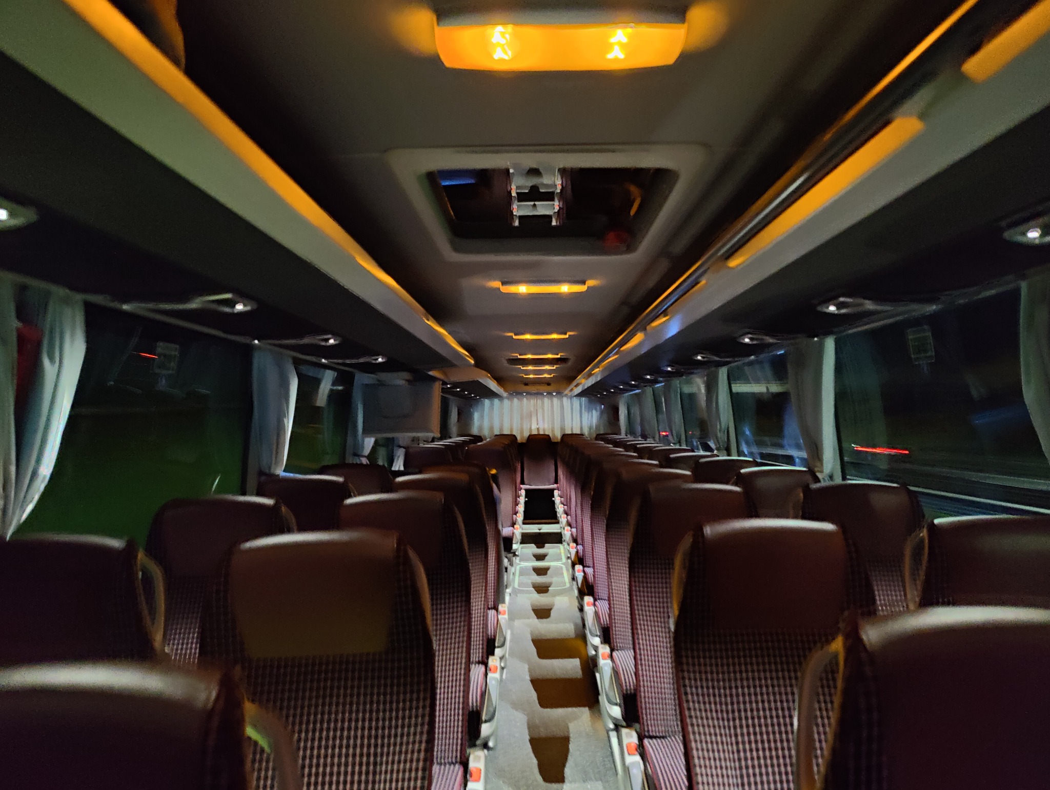 Wnętrze autobusu z rzędami brązowych foteli w kratę, oświetlonych pomarańczowymi lampami sufitowymi, widok z perspektywy pasażera w kierunku tyłu pojazdu.