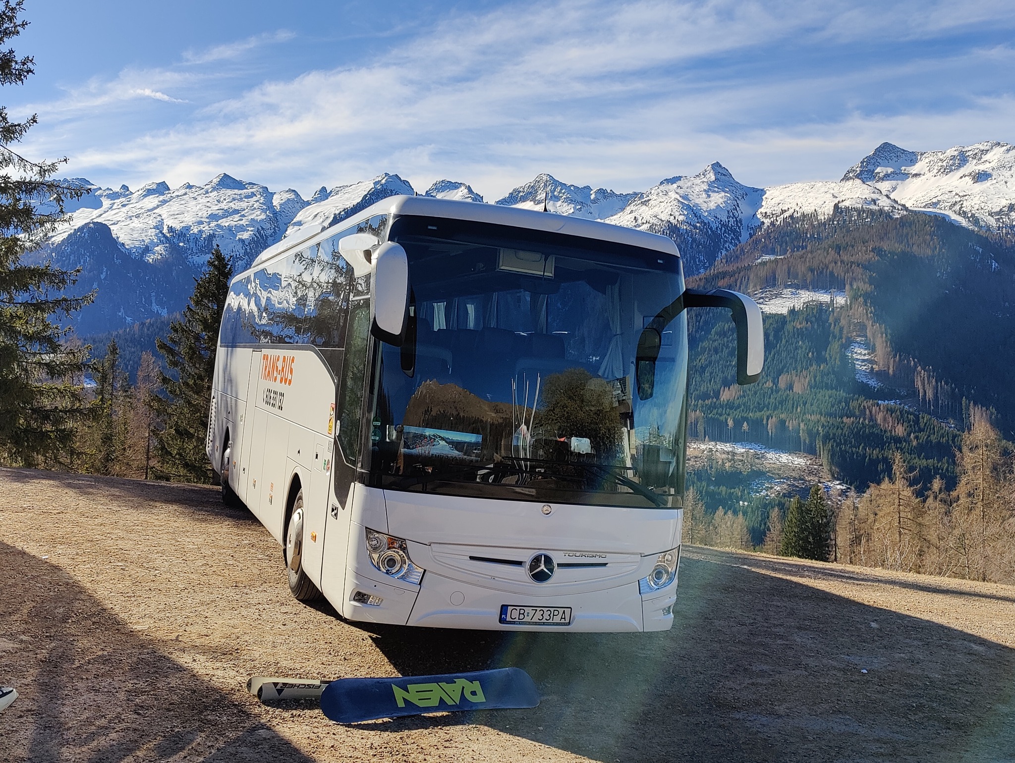 Biały autokar turystyczny z pomarańczowym napisem TRANS-BUS na tle górskich szczytów pokrytych śniegiem i lasów iglastych. Na ziemi leżą narty.
