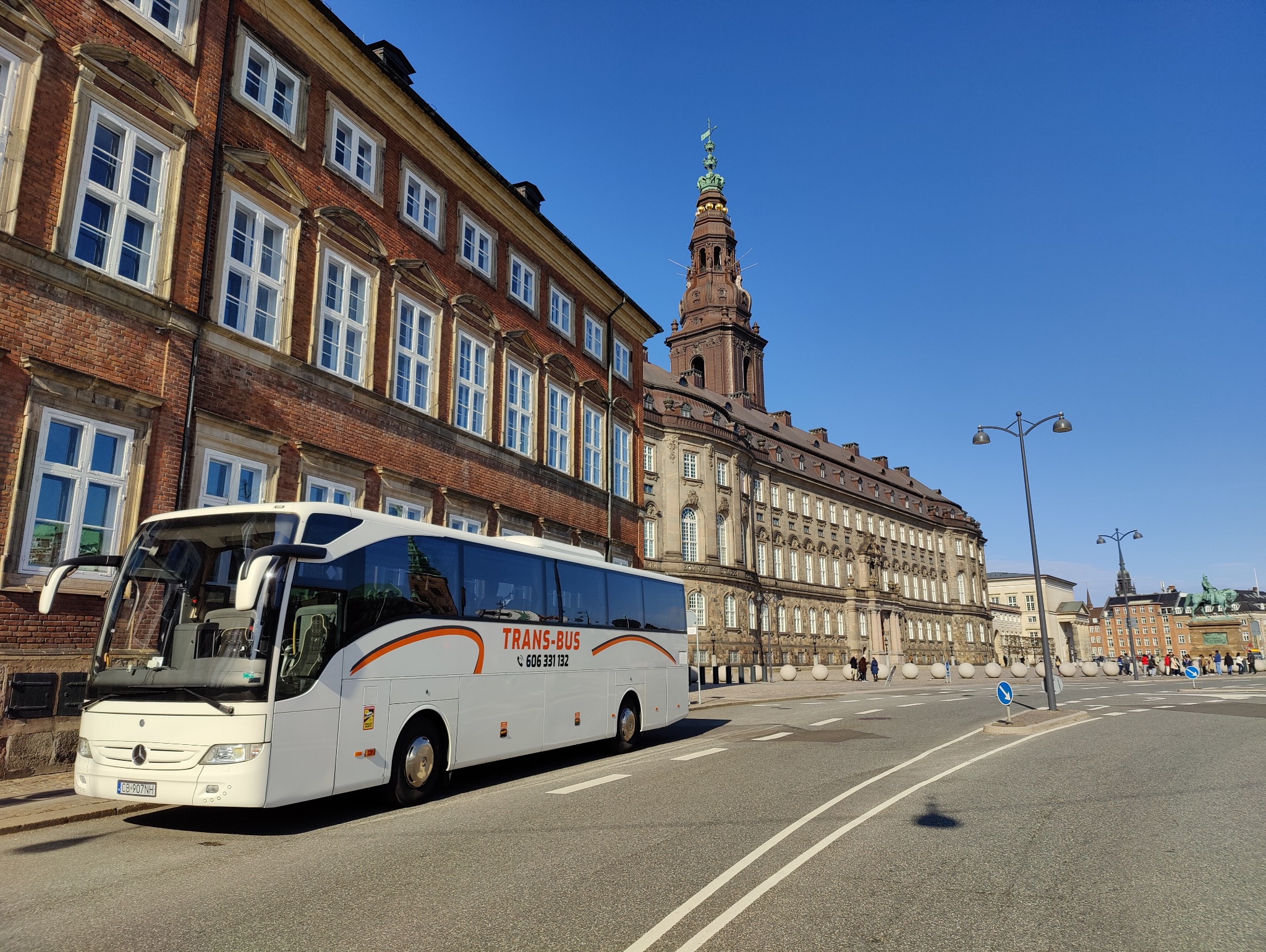 Biały autokar TRANS-BUS zaparkowany na ulicy w Kopenhadze, w tle budynki Christiansborg Palace i Børsen.