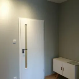 Nowoczesny przedpokój z białą ścianą, drzwiami z przeszkleniem, podłogą z paneli i białą szafką na drewnianych nóżkach, oświetlony listwą z czterema reflektorami.