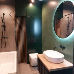 Nowoczesna łazienka z zielonymi płytkami, okrągłym lustrem z podświetleniem LED, białą umywalką nablatową na drewnianym blacie z czarnymi lamelami, czarną armaturą i kabiną prysznicową z deszczownicą.