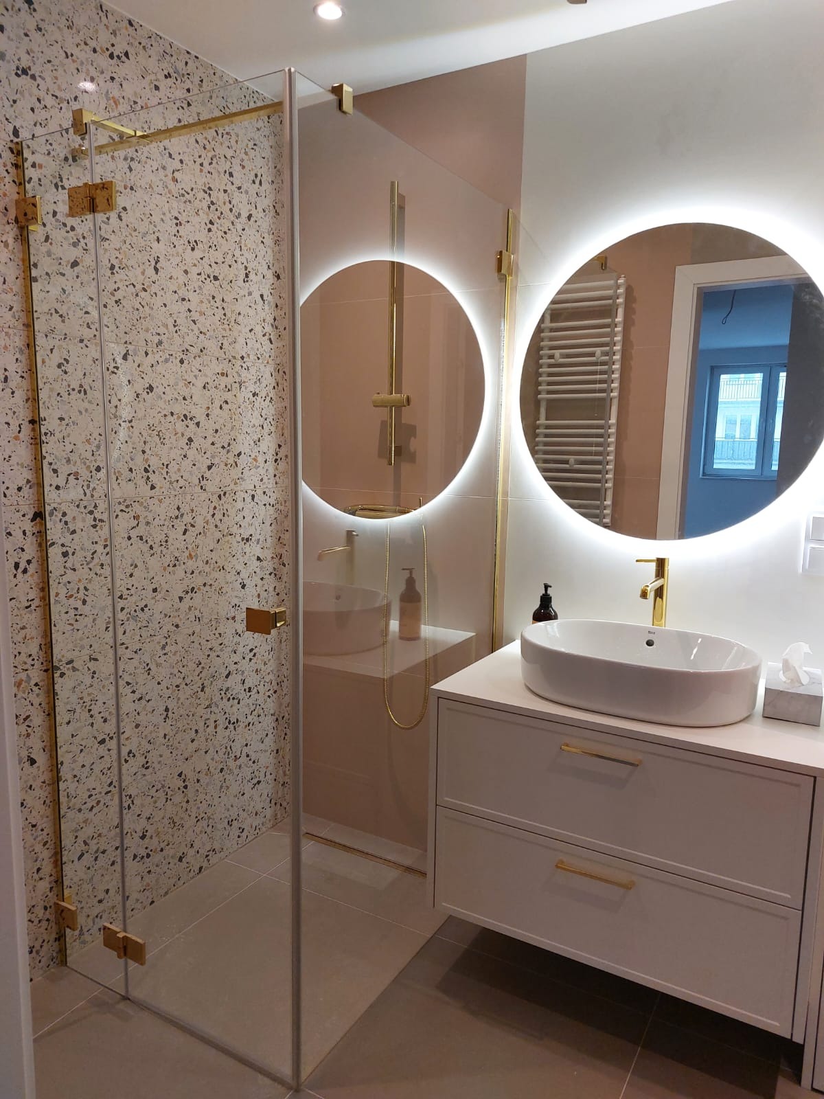 Nowoczesna łazienka z kabiną prysznicową ze złotymi okuciami, umywalką nablatową na szafce z szufladami i okrągłymi lustrami z podświetleniem LED.