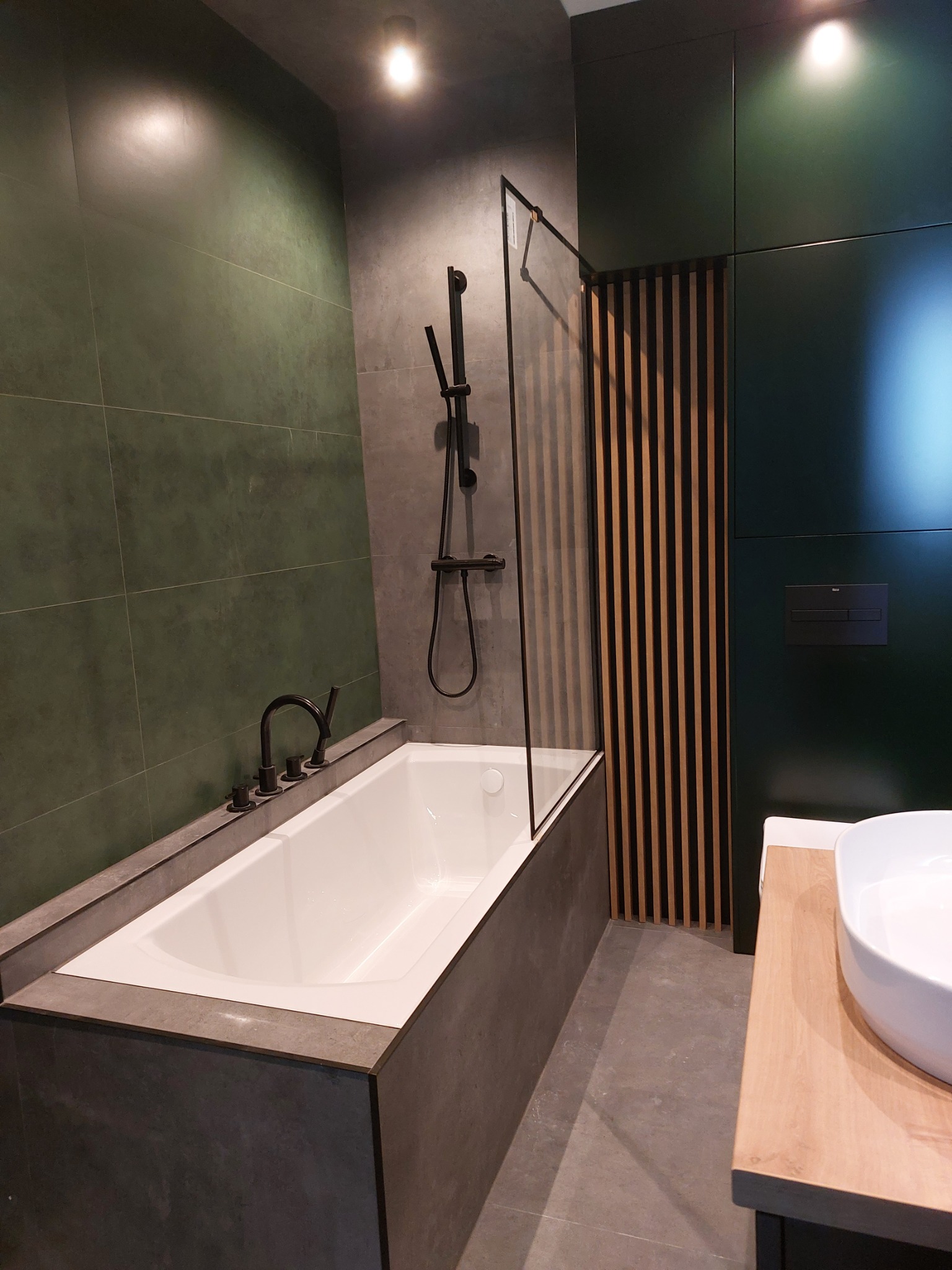 Nowoczesna łazienka z wanną obudowaną szarym kamieniem, czarną armaturą i zielonymi płytkami na ścianie, uzupełniona drewnianymi lamelami i jasnym blatem z umywalką.