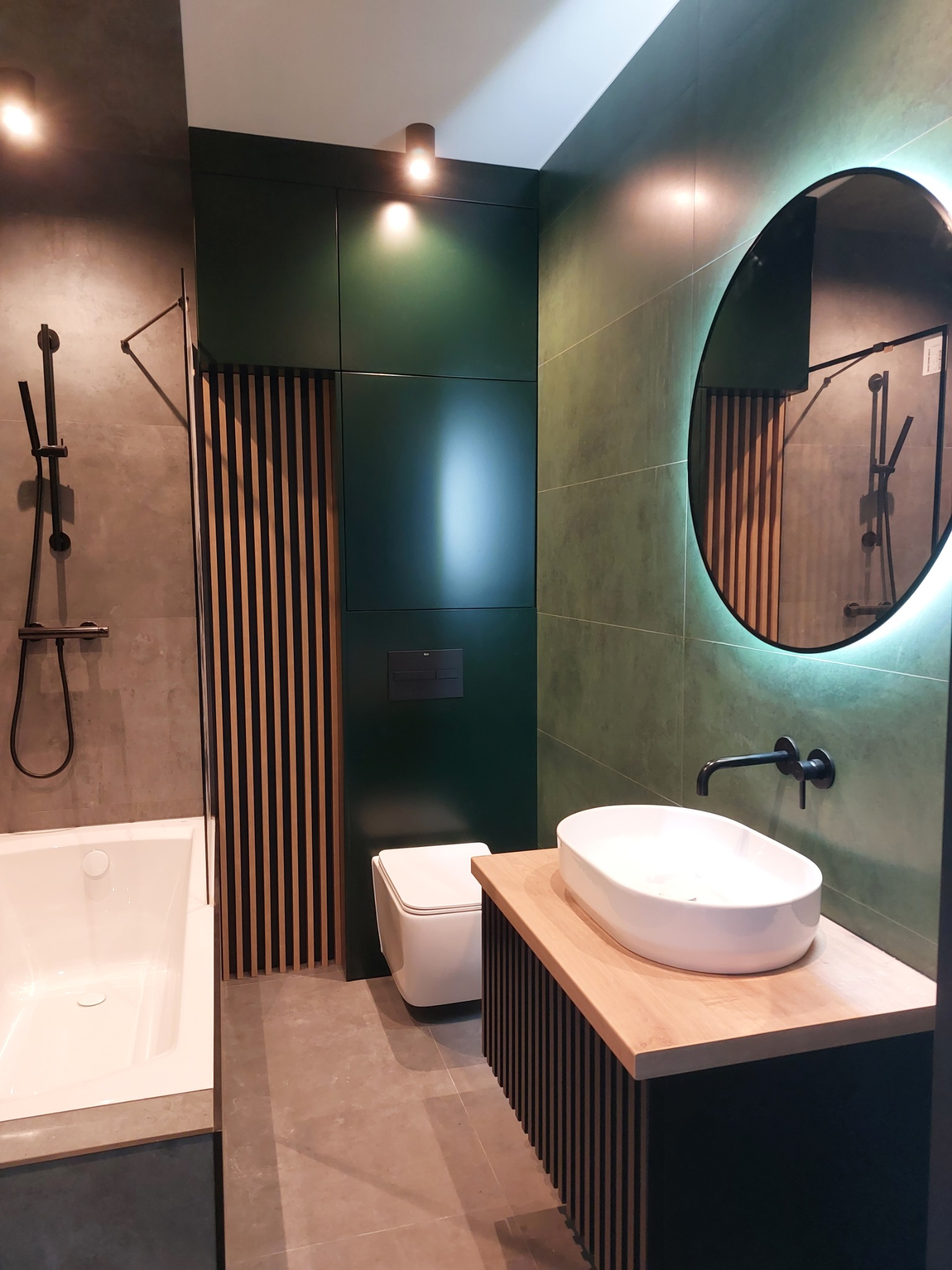 Nowoczesna łazienka z zielonymi płytkami, okrągłym lustrem z podświetleniem LED, białą umywalką nablatową na drewnianym blacie z czarnymi lamelami, czarną armaturą i kabiną prysznicową z deszczownicą.