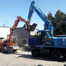 Dwie koparki kołowe Komatsu, jedna pomarańczowa, druga niebieska, stoją na placu budowy w słoneczny dzień, z widocznymi budynkami w tle.