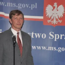 Mężczyzna w szarym garniturze przemawia do mikrofonu na tle ściany z godłem Polski i adresem strony internetowej Ministerstwa Spraw Zagranicznych.