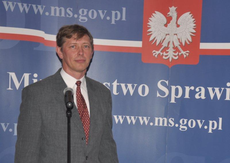 Mężczyzna w szarym garniturze przemawia do mikrofonu na tle ściany z godłem Polski i adresem strony internetowej Ministerstwa Spraw Zagranicznych.