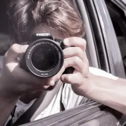 Osoba z aparatem fotograficznym Canon, wystająca przez okno samochodu, z widoczną soczewką obiektywu i fragmentem pasa bezpieczeństwa.