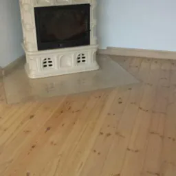 Świeżo wycyklinowana, jasna drewniana podłoga z widocznym piecem kaflowym i fragmentem ściany z cegły, zabezpieczona folią ochronną.