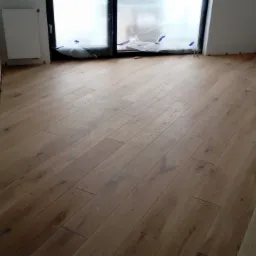 Nowo ułożona, jasna, drewniana podłoga z paneli w pokoju, z zabezpieczonym folią oknem i grzejnikiem na ścianie.