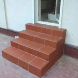 Nowe, ceramiczne schody wejściowe w odcieniach terakoty, prowadzące do otwartych drzwi wejściowych. Widoczne fugi i dopasowana kolorystyka do chodnika.