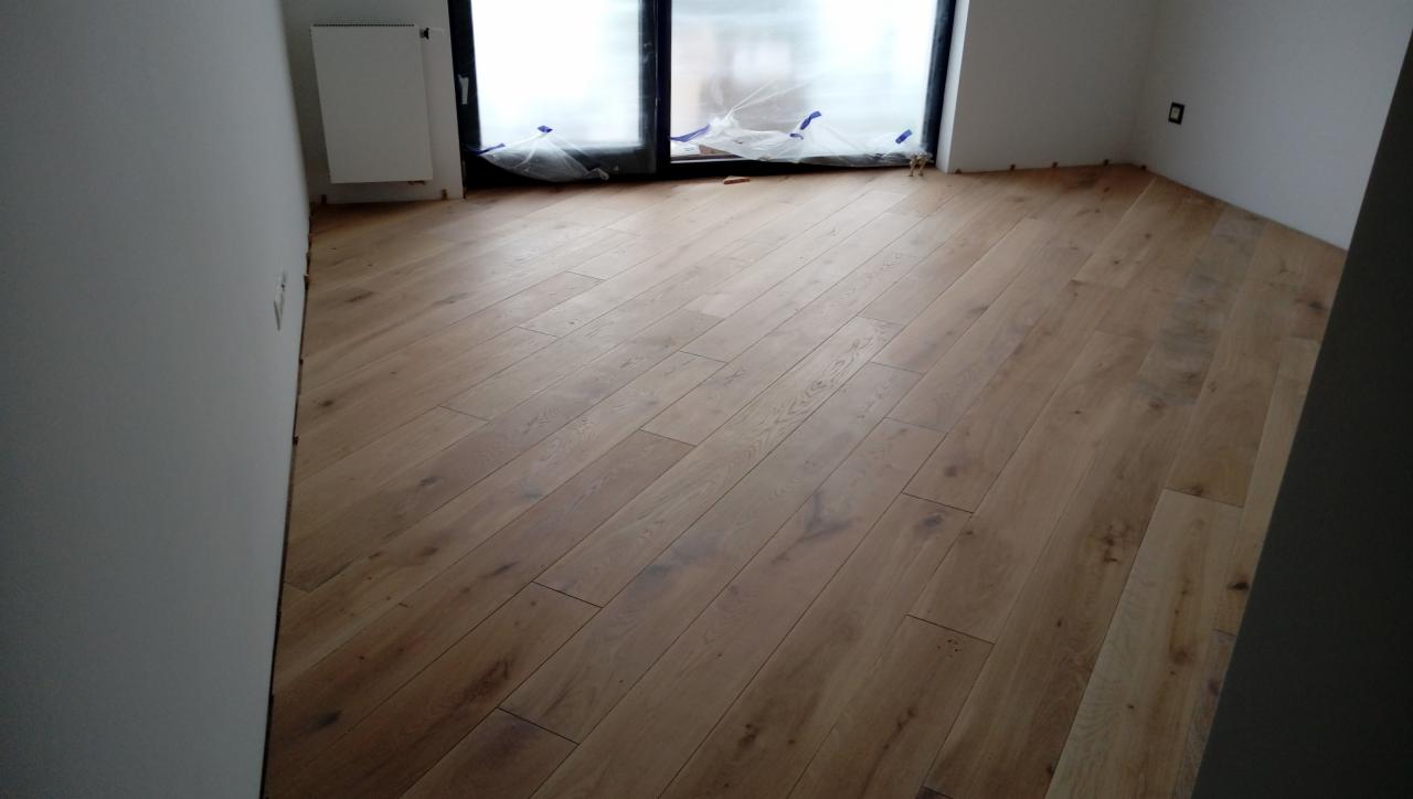 Nowo ułożona, jasna, drewniana podłoga z paneli w pokoju, z zabezpieczonym folią oknem i grzejnikiem na ścianie.