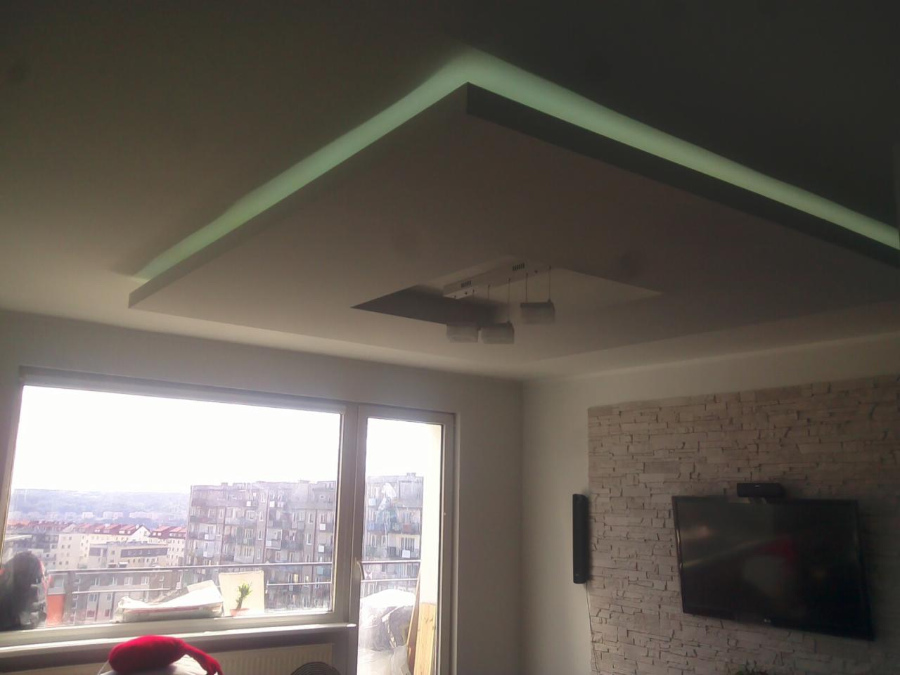 Nowoczesny salon z podwieszanym sufitem z oświetleniem LED, telewizorem na ścianie z kamienia dekoracyjnego i widokiem na miasto z okna.