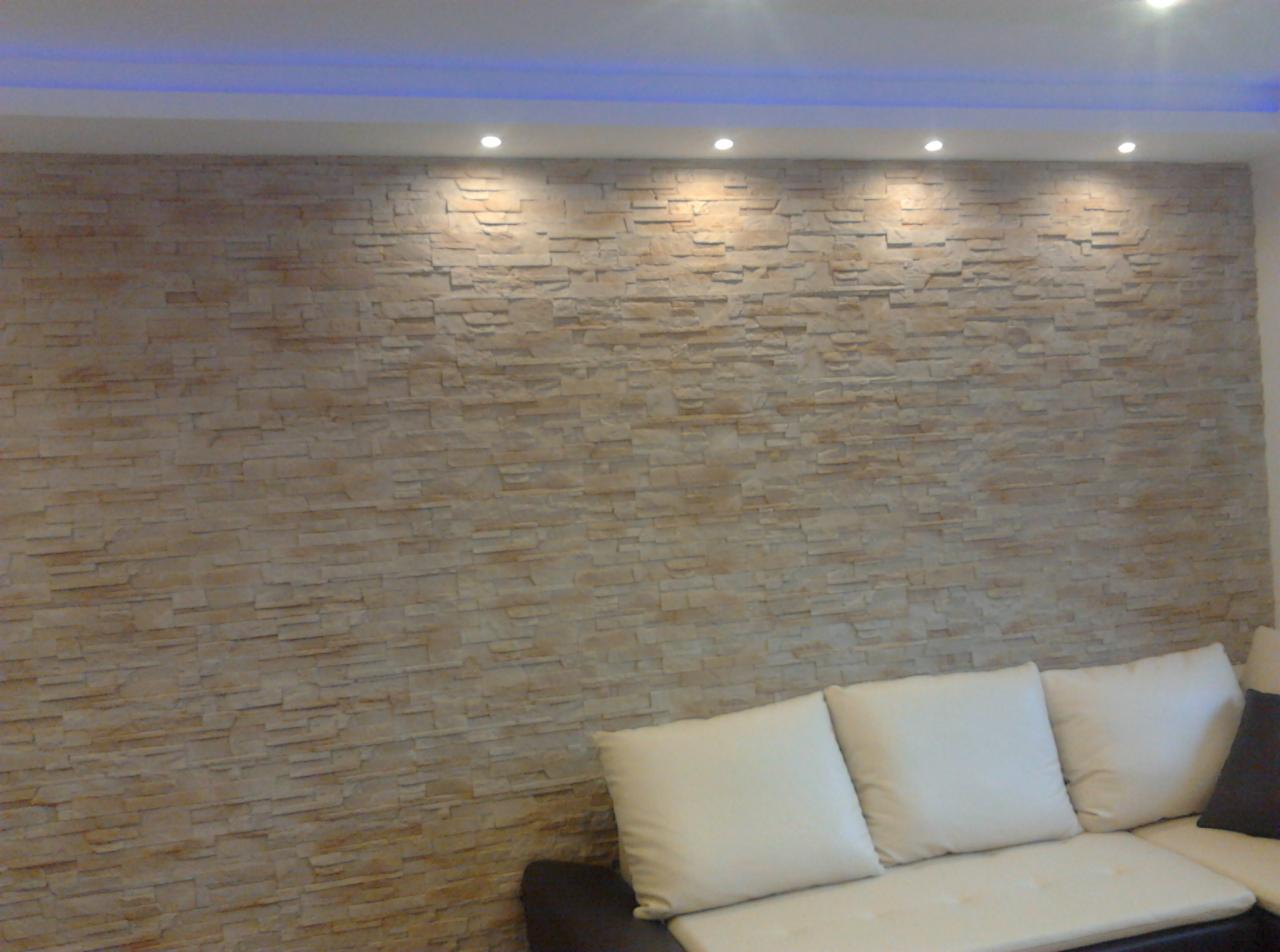 Ściana wyłożona dekoracyjnym kamieniem w odcieniach beżu, oświetlona punktowymi lampami sufitowymi i listwą LED, częściowo widoczna sofa z jasnymi poduszkami.