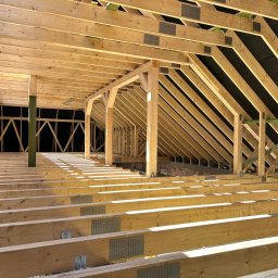 ESP COMFORT - Wnętrze w trakcie budowy: drewniana konstrukcja szkieletowa dachu i podłogi, widoczne elementy mocujące i membrany izolacyjne, oświetlone promieniami słonecznymi wpadającymi przez otwory.