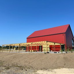 ESP COMFORT - Nowoczesny, czerwony budynek z metalowym dachem i drewnianą pergolą w trakcie budowy, widok z poziomu gruntu na tle błękitnego nieba.