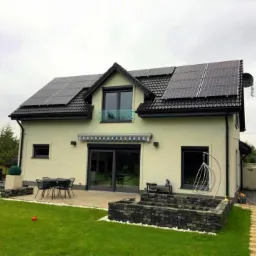 Nowoczesny dom jednorodzinny z panelami słonecznymi na dachu, tarasem z meblami ogrodowymi i ozdobnym oczkiem wodnym z kamienia.