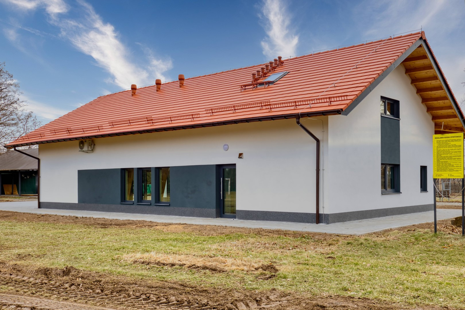 Gotowy, parterowy budynek z czerwonym dachem, białymi ścianami i szarym pasem u dołu, widoczny system rynnowy, okna i drzwi w ciemnej ramie, oraz tablica informacyjna na zewnątrz.