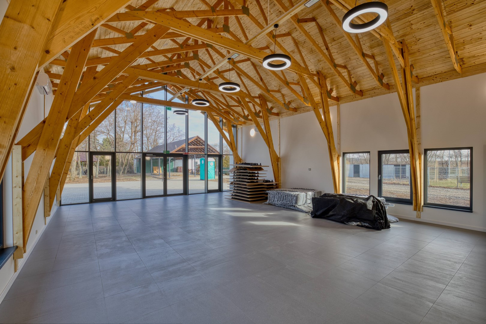 Wnętrze przestronnego budynku z drewnianą konstrukcją dachu, dużymi oknami i szarą podłogą z płytek. Widoczne elementy konstrukcyjne, okrągłe lampy sufitowe i materiały budowlane pod ścianą.