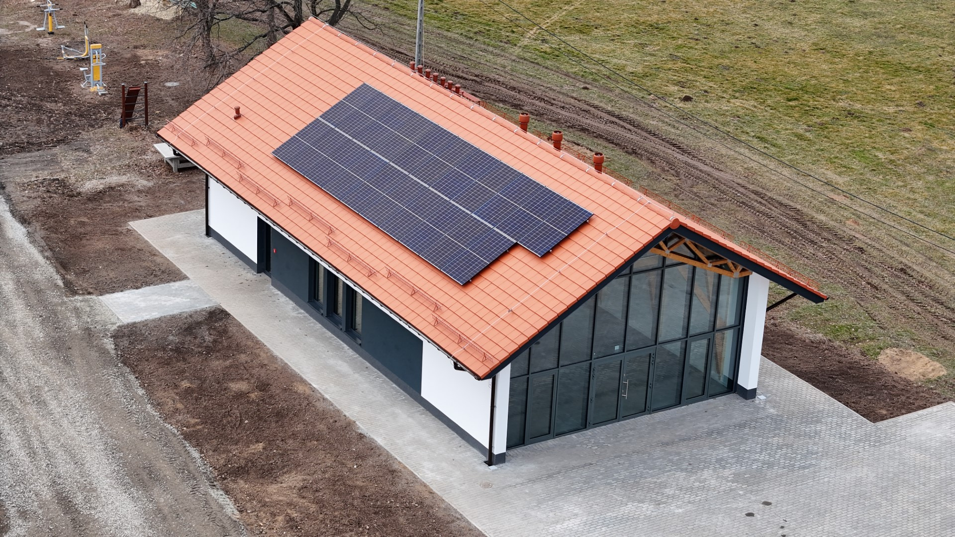 Nowoczesny dom szkieletowy z pomarańczową dachówką i panelami słonecznymi, widok z góry, teren wokół częściowo zagospodarowany kostką brukową i ziemią.
