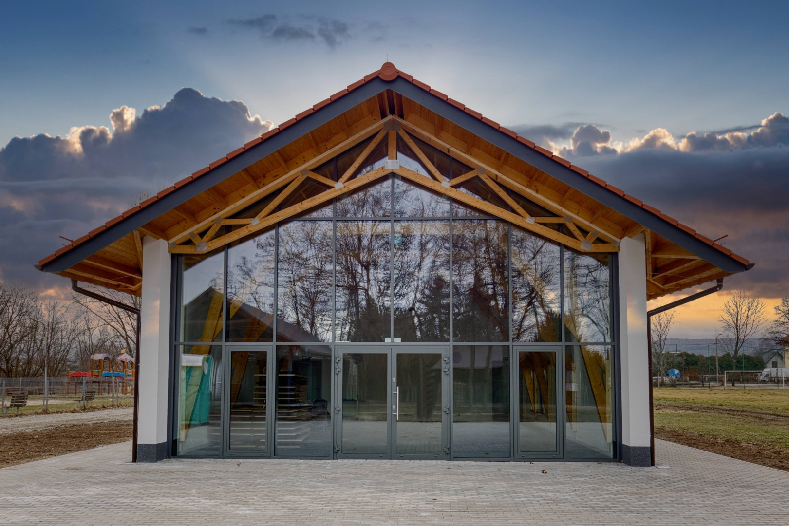 Nowoczesny budynek z drewnianą konstrukcją dachu i przeszkloną fasadą, odbijającą drzewa i zachmurzone niebo o zmierzchu, na pierwszym planie bruk.